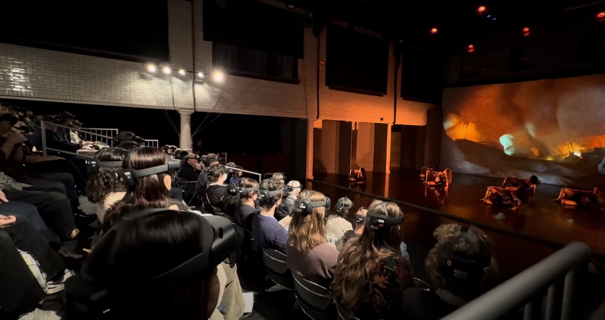 A dancer mid-leap surrounded by flowing holographic light trails and geometric shapes that respond to their movement. Audience members in headsets visible in the background.