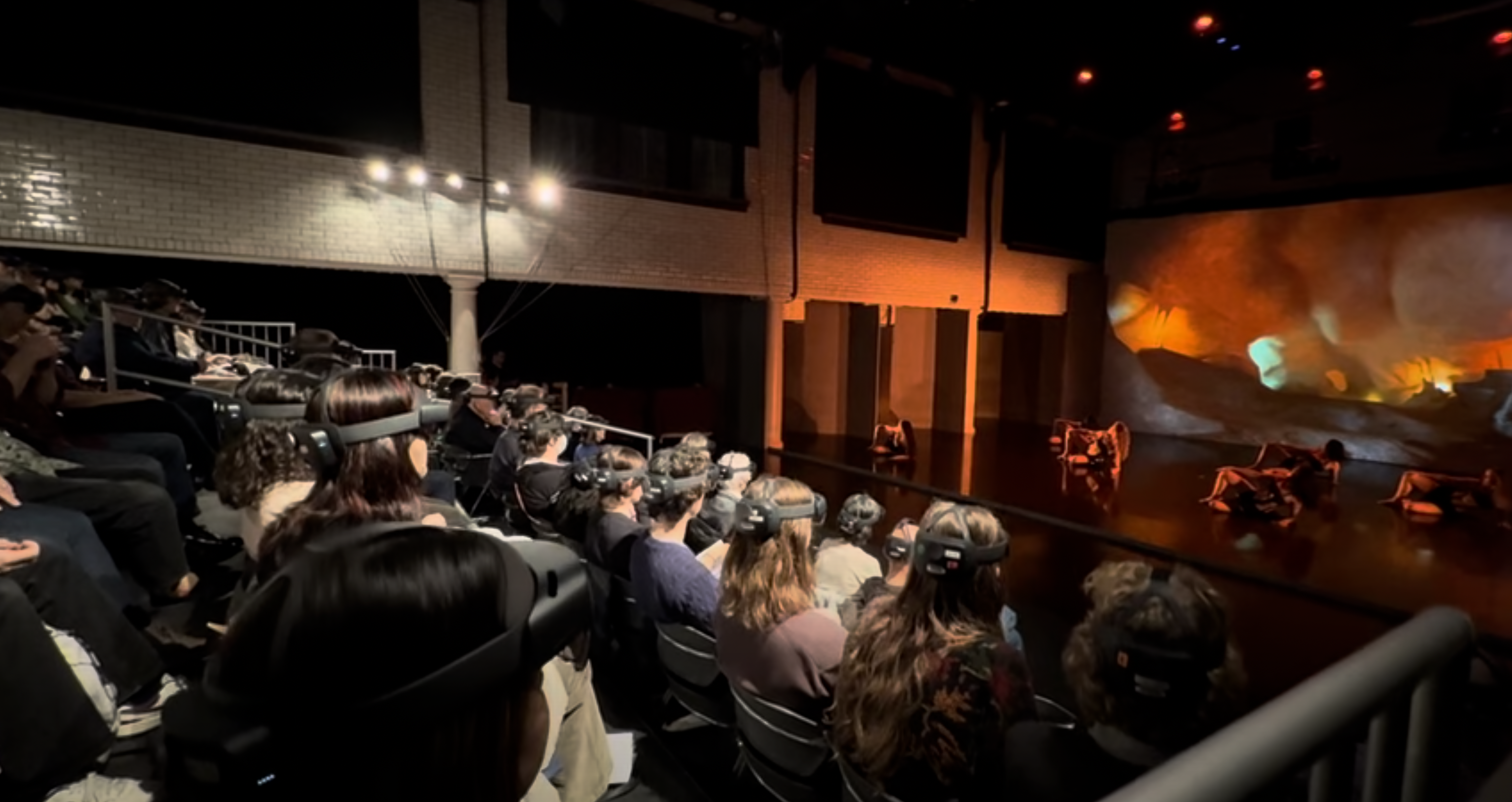 A dancer mid-leap surrounded by flowing holographic light trails and geometric shapes that respond to their movement. Audience members in headsets visible in the background.
